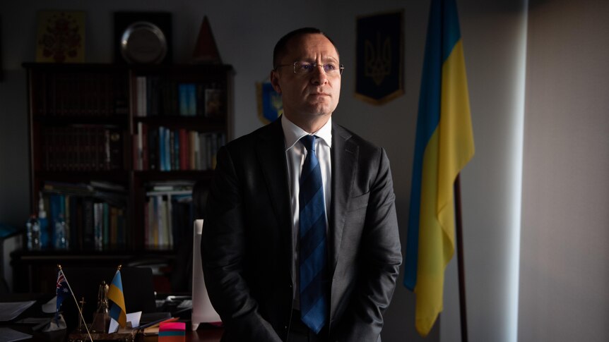 一个短发戴眼镜的中年男子坐在在乌克兰国旗前面的桌子边缘。